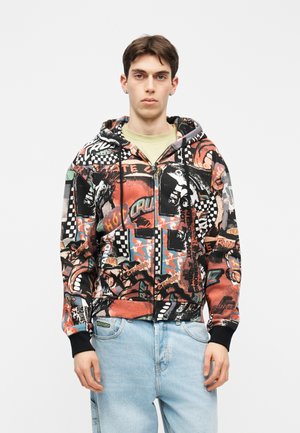 Mladý muž v barevné mikině na zip s grafikou šachovnice a plamenů, ve světlých modrých džínách.