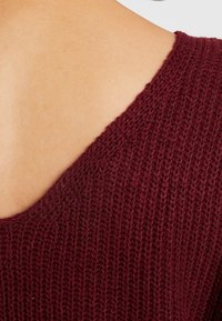 Bordeauxfarbener Strickpullover mit weitem V-Ausschnitt und strukturiertem Material, der den Rücken zeigt, mit einem gerippten Saum.