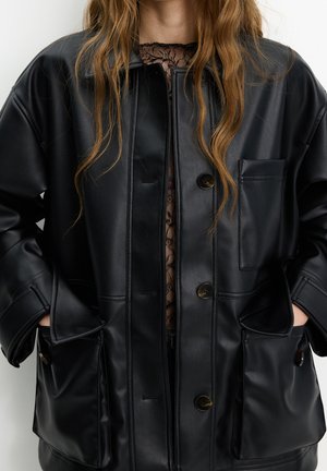 Femme portant une veste en cuir noire boutonnée sur un top en dentelle noire transparente, avec de longs cheveux lâches ondulés et les mains dans les poches de la veste.