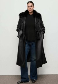 Lungo cappotto di pelle nera con colletto in pelliccia, vita con lacci e tasche laterali. Abbinato a un maglione a coste nero e a jeans in denim scuro a zampa d'elefante.