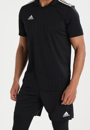 Mann trägt schwarze Adidas-Sportkleidung mit weißem Logo auf der Brust und Shorts, steht mit entspannt an den Seiten hängenden Armen.