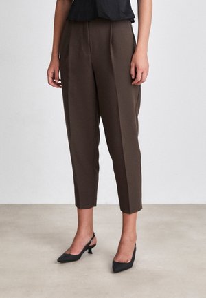 Femme portant un pantalon marron coupé sur mesure et des talons slingback noirs, debout sur un sol clair contre un mur blanc.
