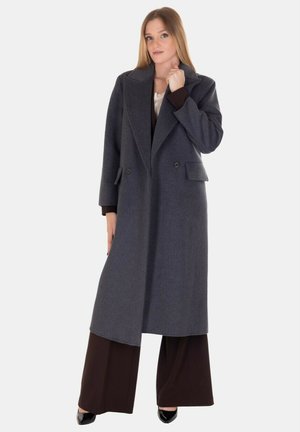 Cappotto di lana grigio con design doppiopetto, ampi revers e tasche laterali, indossato su pantaloni larghi marroni. Tessuto liscio e vestibilità lunga.