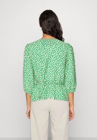Blouse verte à basque avec imprimé floral, manches trois-quarts et encolure ronde. Tissu léger avec une surface texturée.