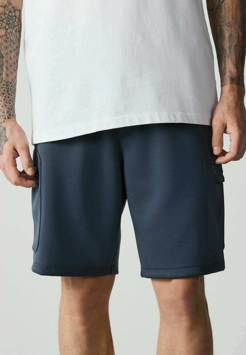 Homme portant un short cargo bleu marine et une chemise blanche, montrant les avant-bras tatoués et les mains reposant près des poches.