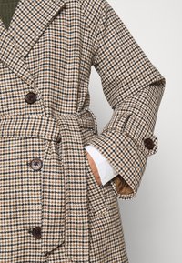 Cappotto a quadri marrone e bianco con ampio colletto, bottoni doppi, cintura a nastro e maniche con polsini. Tessuto morbido e testurizzato.