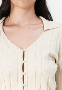 Gebreid vest in lichtbeige met een V-hals, voorzien van een kabelpatroon en vijf parelknoopjes langs de voorkant. Effen textuur.