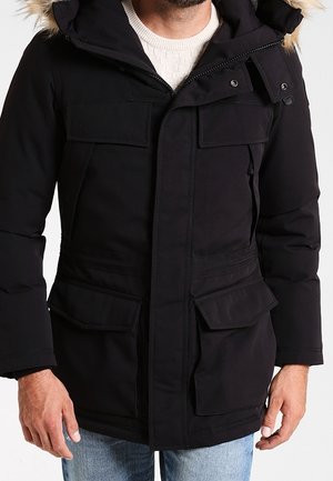Mann trägt eine schwarze Winterparka mit pelzbesetzter Kapuze und mehreren Fronttaschen, kombiniert mit hellblauen Jeans und weißem Pullover.