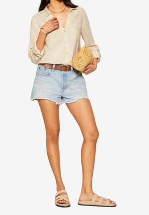 Kobieta w beżowej koszuli na guziki, jasnych, postrzępionych szortach z denimu, brązowym pasku, beżowych sandałach, trzymająca plecioną kopertówkę.