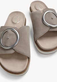 Sandales plates en daim taupe avec larges brides croisées et grande boucle circulaire argentée sur une semelle blanche.