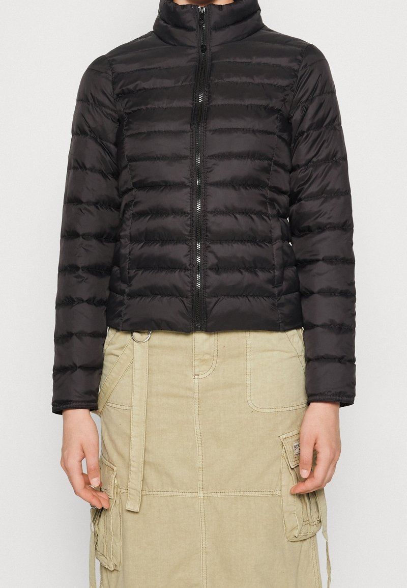 Veste noire matelassée à fermeture éclair portée avec une jupe cargo beige dotée de poches et de détails de sangles sur une personne debout.