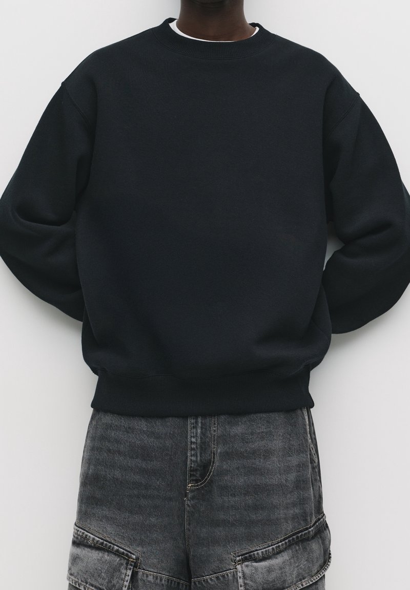 Personne portant un sweat-shirt noir uni à col rond et un short en denim gris foncé avec des poches à rabat devant sur un fond clair.