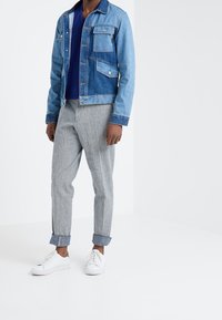 Jeansjacke in hellblau mit mehreren Taschen, getragen über einem marineblauen V-Ausschnitt-Shirt, kombiniert mit grauen gestreiften engen Hosen und weißen Sneakers.