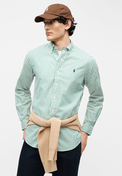 Νεαρός άνδρας που φορά καφέ καπέλο, πουκάμισο με πράσινες και άσπρες ρίγες και κουμπιά, μπλε παντελόνι, με μπεζ πουλόβερ δεμένο γύρω από τη μέση του, κοιτάζει αριστερά.