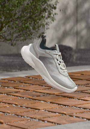 Chaussure de sport gris clair avec texture lisse, tissu respirant, logo sur le côté et semelle blanche, posée sur une surface en bois.