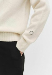 Crème trui van zacht gebreid materiaal, met geribde manchetten en een klein rond logo detail op de linker mouw. Zwarte broek zichtbaar.