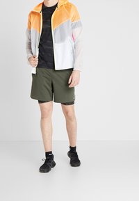 Chaqueta ligera y colorida con paneles en naranja, gris y blanco. Combinada con pantalones cortos verdes oliva y zapatillas negras. Camiseta negra de camuflaje debajo.