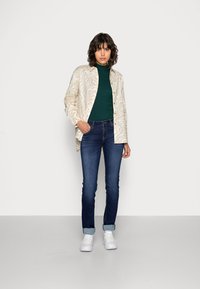 Licht gestreept overhemd in beige en crème, gecombineerd met een donkergroen coltrui, donkerblauwe spijkerbroek en witte sneakers.