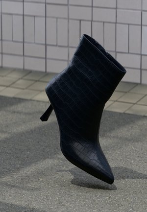 Bottine en cuir noir texturé avec un bout pointu et un talon fin, posée sur un trottoir devant un mur carrelé.