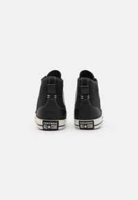 Zapatillas altas de lienzo negro con costuras blancas, suelas de goma y marca All Star en la parte trasera, que incluyen un detalle de logotipo circular.
