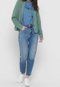 Ljusgrön kavaj över en denimskjorta med volangdetaljer, kombinerad med ljusblå tapered jeans och vita sneakers. Enkel och avslappnad look.