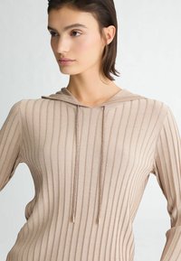 Haut à capuche beige côtelé avec un subtil éclat, présentant un cordon de serrage avec des embouts métalliques et une silhouette ajustée. Détail en tissu texturé visible.