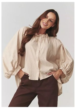 Beżowa bluzka z guzikami, z falbaniastym dekoltem i dużymi, puffowymi rękawami. Teksturowany materiał; luźny krój. Zestawiona z brązowymi spodniami.