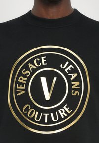 Črna trenirka z velikim zlatim okroglim logotipom, ki vsebuje napis "VERSACE JEANS COUTURE" in reliefno črko "V" v središču.