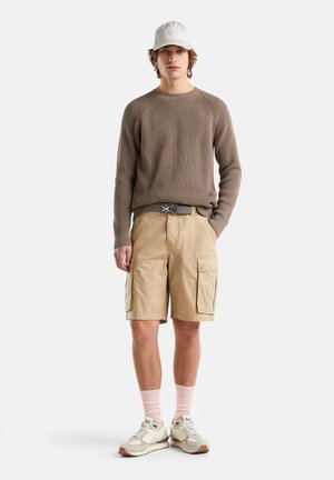 Suéter marrón texturizado, pantalones cortos cargo color caqui con bolsillos laterales, cinturón gris, calcetines de color rosa claro y zapatillas beiges. Lleva una gorra suave con un logotipo en la cabeza.