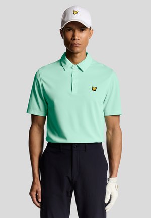 Mintgrøn polo-shirt med krave og gult broderet logo, parret med mørke bukser og en hvid kasket. Inkluderer en golfhandske på venstre hånd.