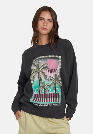 Vrouw draagt een zwart sweatshirt met een kleurrijke palmboom, oceaangolven en zonsondergangafbeelding, gecombineerd met beige broek.