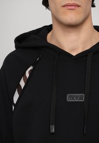 Hoodie noir avec un design en découpe graphique, capuche à lacets avec cordons de serrage noirs, et un patch logo affichant "VERSACE JEANS COUTURE."