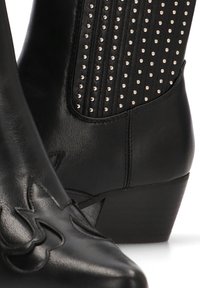 Botas negras de cuero hasta el tobillo con diseños en relieve, detalles de tachuelas y un panel texturizado. Tacón bajo y apilado con una forma de punta afilada.