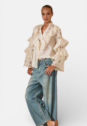 Donna che indossa una camicetta in pizzo color crema con maniche arricciate e jeans blu a gamba larga, in posa con una mano sul fianco su uno sfondo bianco.