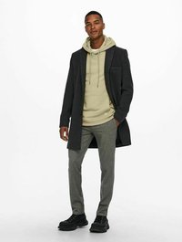 Donkergrijze overjas, lichtgroene hoodie, grijzige patroonbroek en zwarte schoenen. Kenmerken zijn een gestructureerde stof en casual laagjes.
