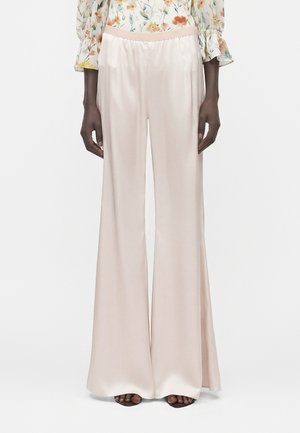 Osoba ubrana w kremowe, jedwabne spodnie o szerokich nogawkach oraz kwiecistą bluzkę z długim rękawem, stojąca z rękami swobodnie opuszczonymi wzdłuż ciała.