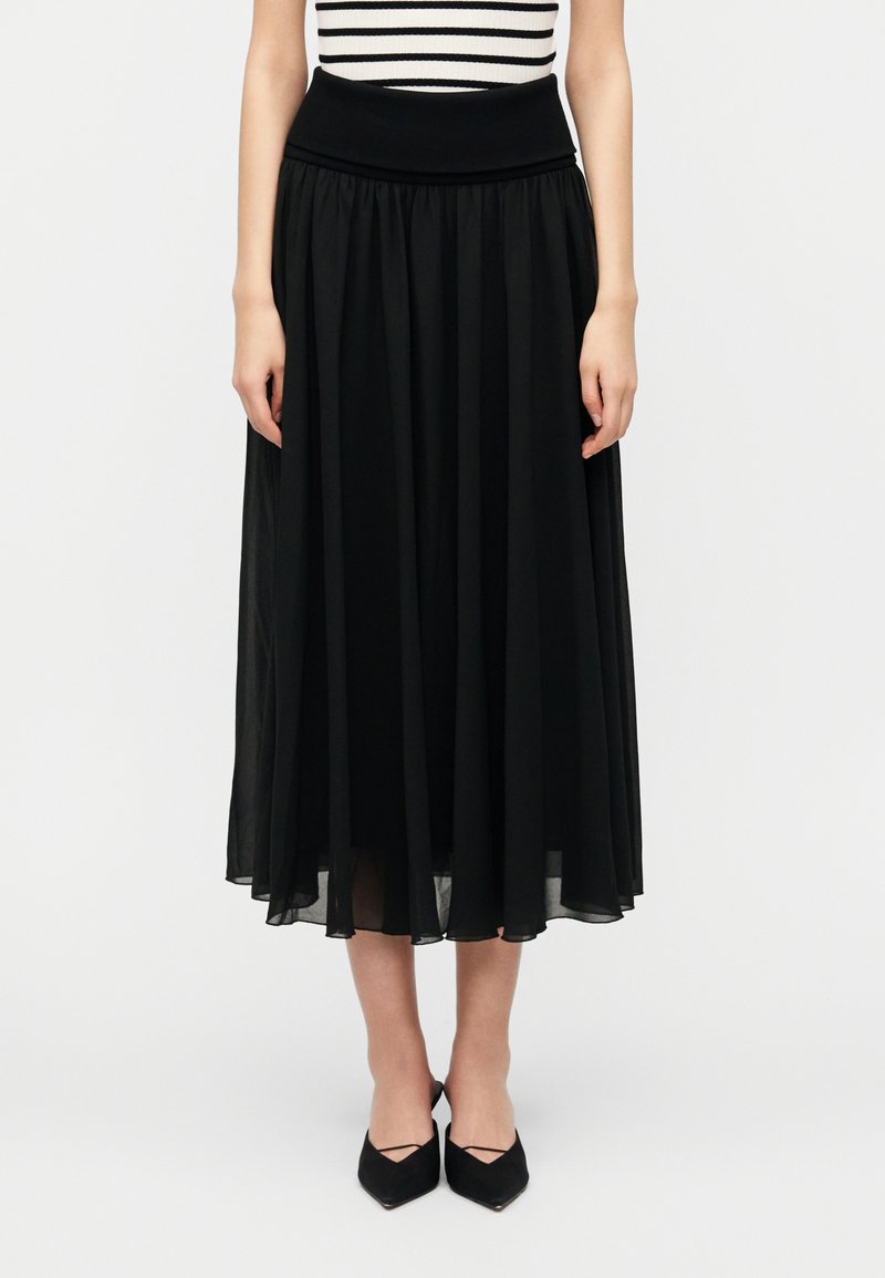 Μακριά μαύρη φούστα από ελαφρύ υλικό με φαρδιά μέση. Διαθέτει απαλές πιέτες, που τελειώνουν λίγο πάνω  από τους αστραγάλους. Φοριέται με μαύρα παπούτσια.