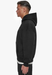 Schwarze Kapuzenjacke mit Kängurutasche, gerippten Bündchen und einer Kordelzugkapuze. Glatte Textur, lässiges Design und ein einfaches, gesticktes Logodetail.