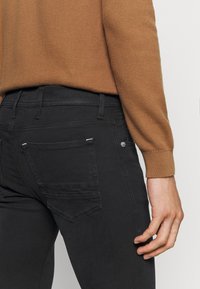 Person som bär svarta jeans och en brun långärmad stickad tröja, med höger hand avslappnad vid sin sida.