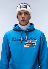 Felpa con cappuccio blu con logo e stampa grafica; presenta un collo con coulisse, abbinata a un berretto di lana bianco con design a bandiera.