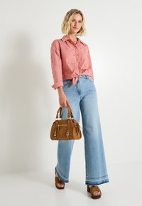 Femme portant un blouse brodé rose, un jean large bleu clair, des sandales à talons marron et tenant un sac à main marron.