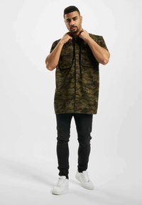 Giacca camuffata a maniche corte, con due tasche frontali e motivo testuale; abbinata a jeans neri e sneakers bianche.