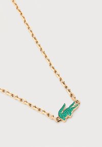 Χρυσό κολιέ με αλυσίδα και μενταγιόν σε σχήμα πράσινου αλιγάτορα διακοσμημένο με μικρές πράσινες πέτρες σε απλό ανοιχτό φόντο.