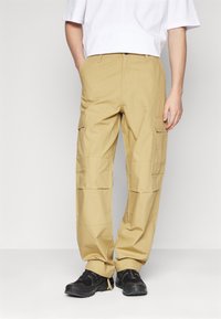 Beige cargo bukser med flere lommer, en afslappet pasform og manchetter, parret med sorte sko og en hvid kortærmet skjorte.