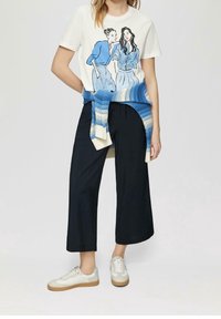 Bílá grafická tričko s ilustracemi, černé široké kalhoty a krémové tenisky s béžovou podrážkou; modrý pletený šátek uvázaný v pase.