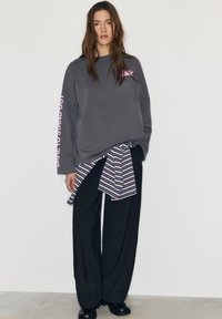 Szürke, túlméretezett pulóver rózsaszín nyomattal az ujjon, fekete bő szárú nadrággal és kék csíkos megkötős inggel párosítva. Sima textúrák.