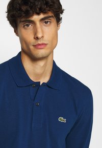 Marineblaue Polohemd aus strukturiertem Stoff, mit einer Knopfleiste mit zwei Knöpfen und einem kleinen grünen Krokodil-Logo auf der linken Brust.