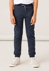 Marineblauwe sweatpants met een trekkoord in de taille, taps toelopende pijpen en ribbenmanchetten bij de enkels, gecombineerd met lichte sneakers met witte strepen.