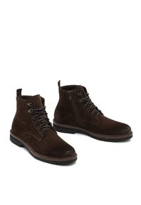 Un paio di stivaletti in suede marrone scuro con lacci neri, cerniera laterale e suola in gomma nera su uno sfondo bianco.