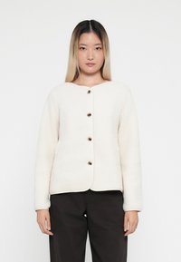 Veste en polaire crème avec un col arrondi, fermeture à boutons sur le devant et manches longues. Pantalon noir porté en dessous.